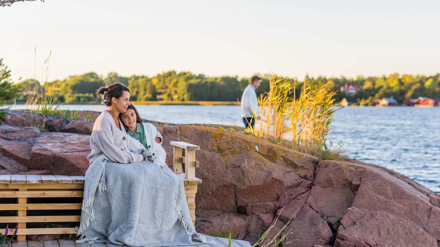 Äiti ja tytär istuvat rantakalliolla Ahvenanmaalla, taustalla meri ja ilta-auringon lempeä valo.