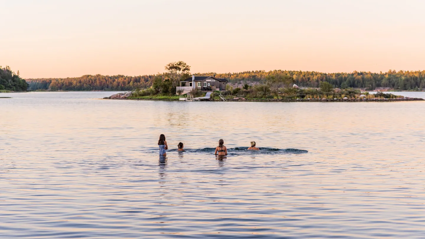 Neljä henkilöä uimassa aurngonlaskun aikaan Ahvenanmaalla, taustalla rauhallinen meri ja lämmin iltavalo.