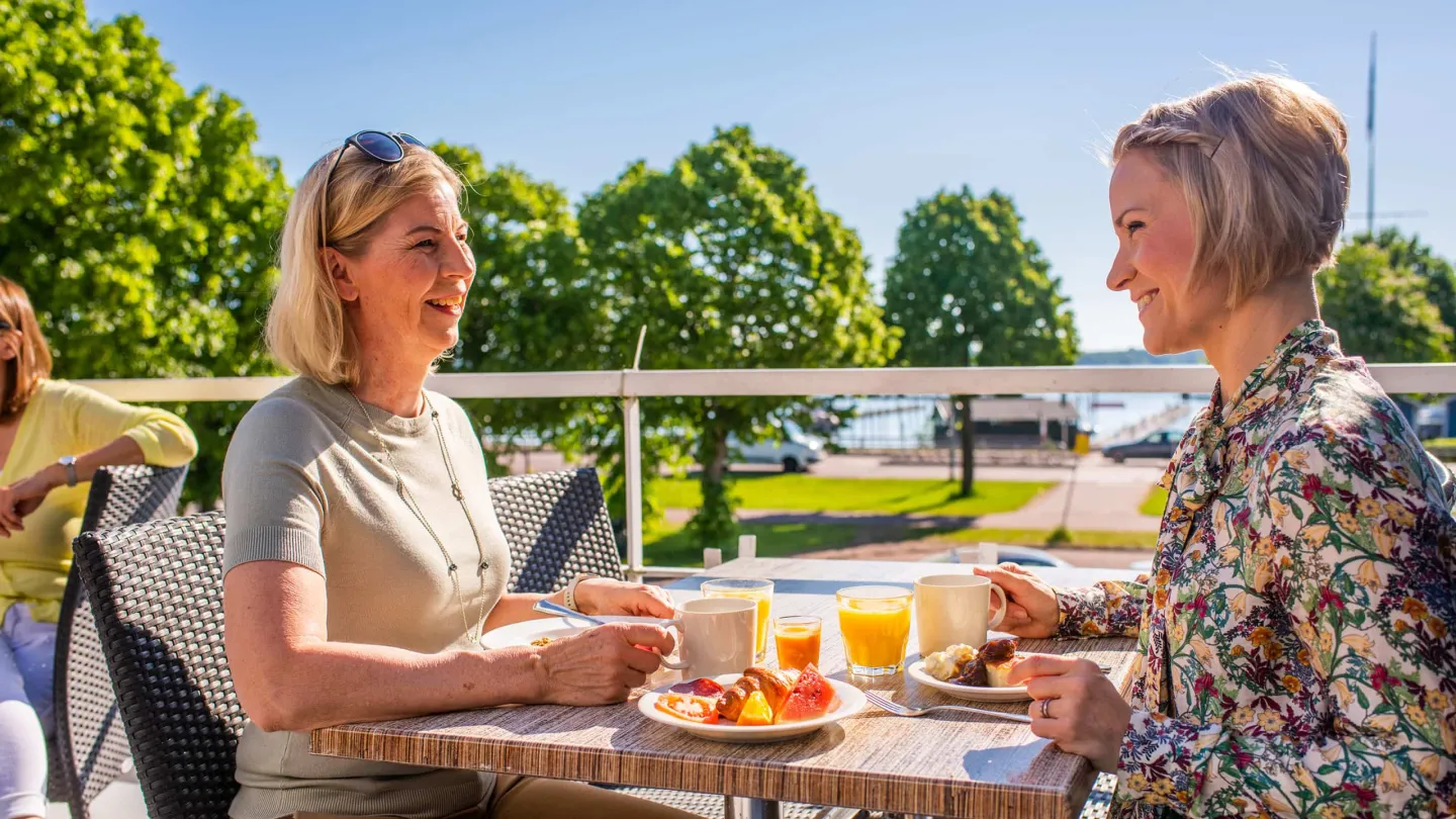 Two women enjoying an outdoor hotel breakfast on a sunny terracce in Mariehamn, Åland. Local ingredients and morning sunlight.