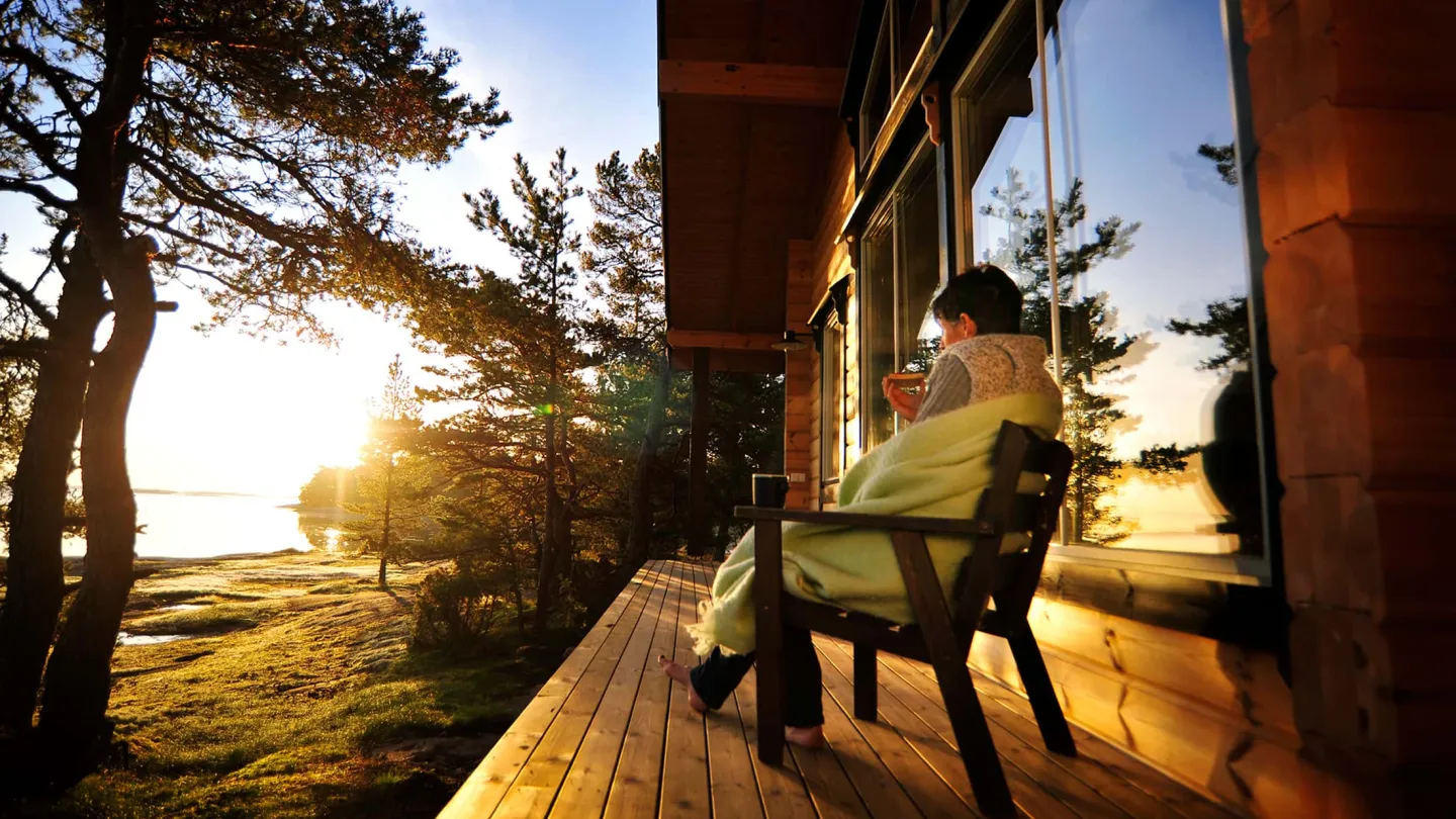 Henkilö istuu viltin alla mökin terassilla Ahvenanmaalla ja juo kavia, taustalla meri ja nouseva aurinko.